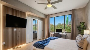 A modern hotel room with a large bed, a desk with a computer, a television mounted on the wall, and a view of greenery outside. - Modern 2BR/2BA Retreat • Pool • Gym • Free Parking (Phoenix)