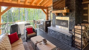 Una terraza de madera acogedora con chimenea de piedra, zona de estar y vistas al bosque.