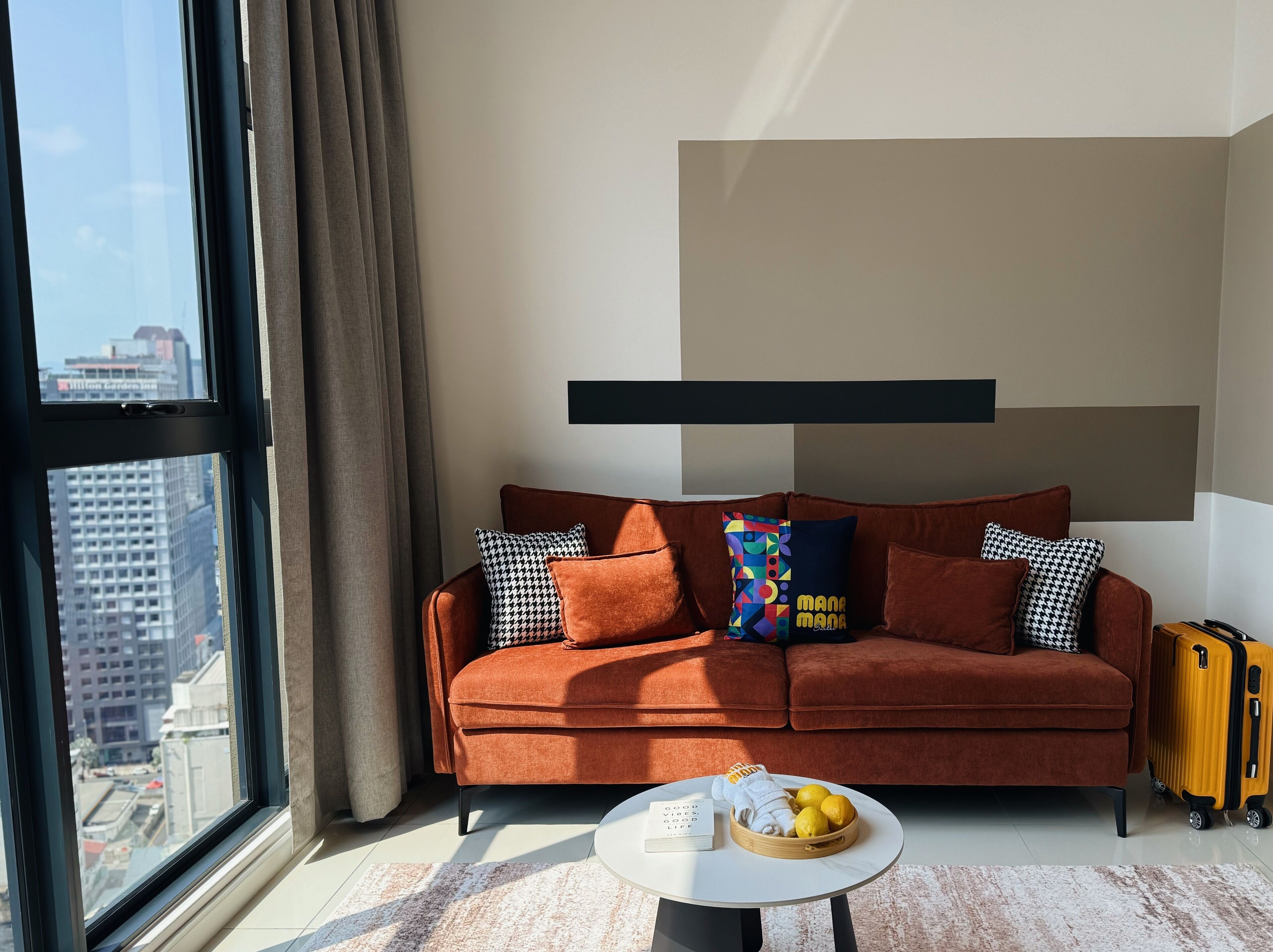 Ein modernes Wohnzimmer mit einem braunen Sofa, gemusterten Kissen, einem runden Couchtisch mit Fruchtkorb und einem Koffer.