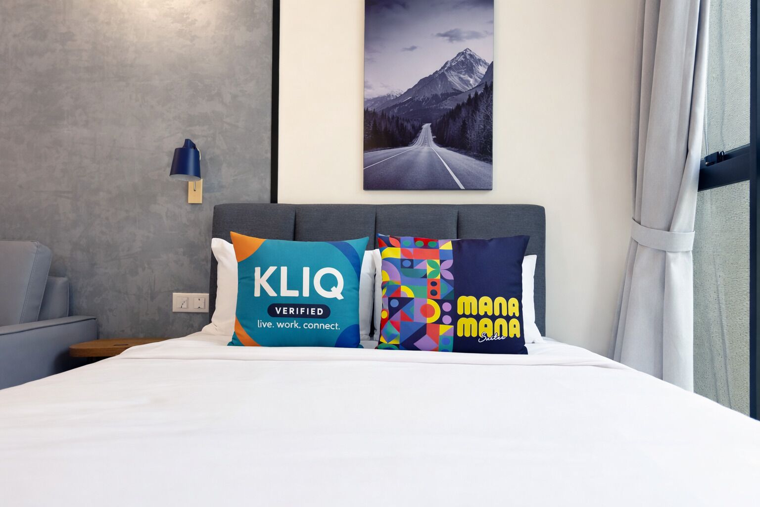 A hotel room with a bed, two pillows with text, a nightstand, a lamp, a window with curtains, and a mountain landscape painting on the wall.