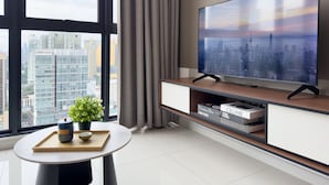 Una sala de estar moderna con un televisor de pantalla plana grande, una mesa redonda con una planta y vistas a un paisaje urbano a través de ventanas amplias.