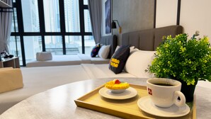 Un salón moderno con un sofá, una mesita de centro con una bandeja que sostiene un postre y una taza de café, y vistas de altos edificios a través de amplios ventanales.