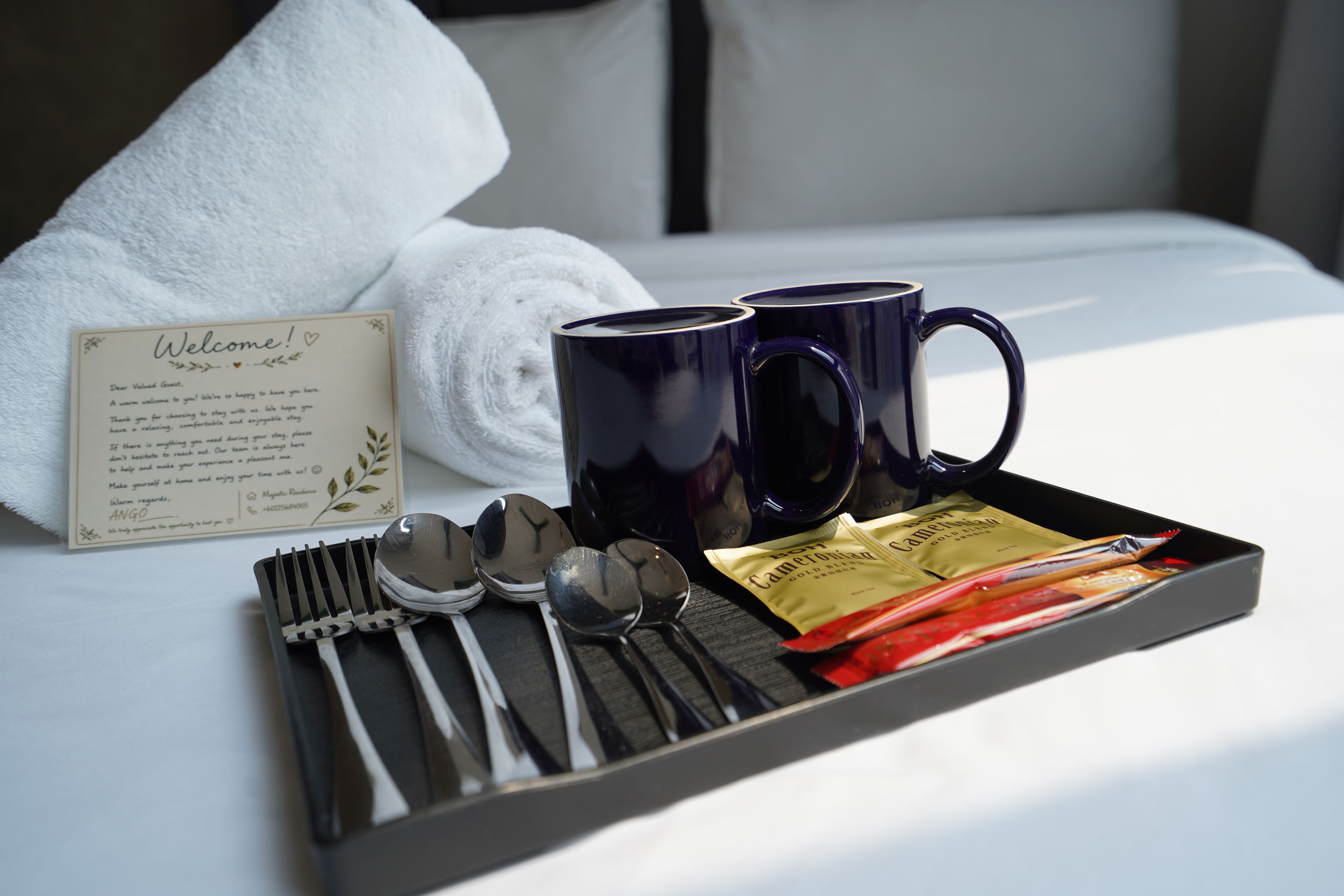 A tray with a welcome card, two mugs, cutlery, and tea packets on a bed.