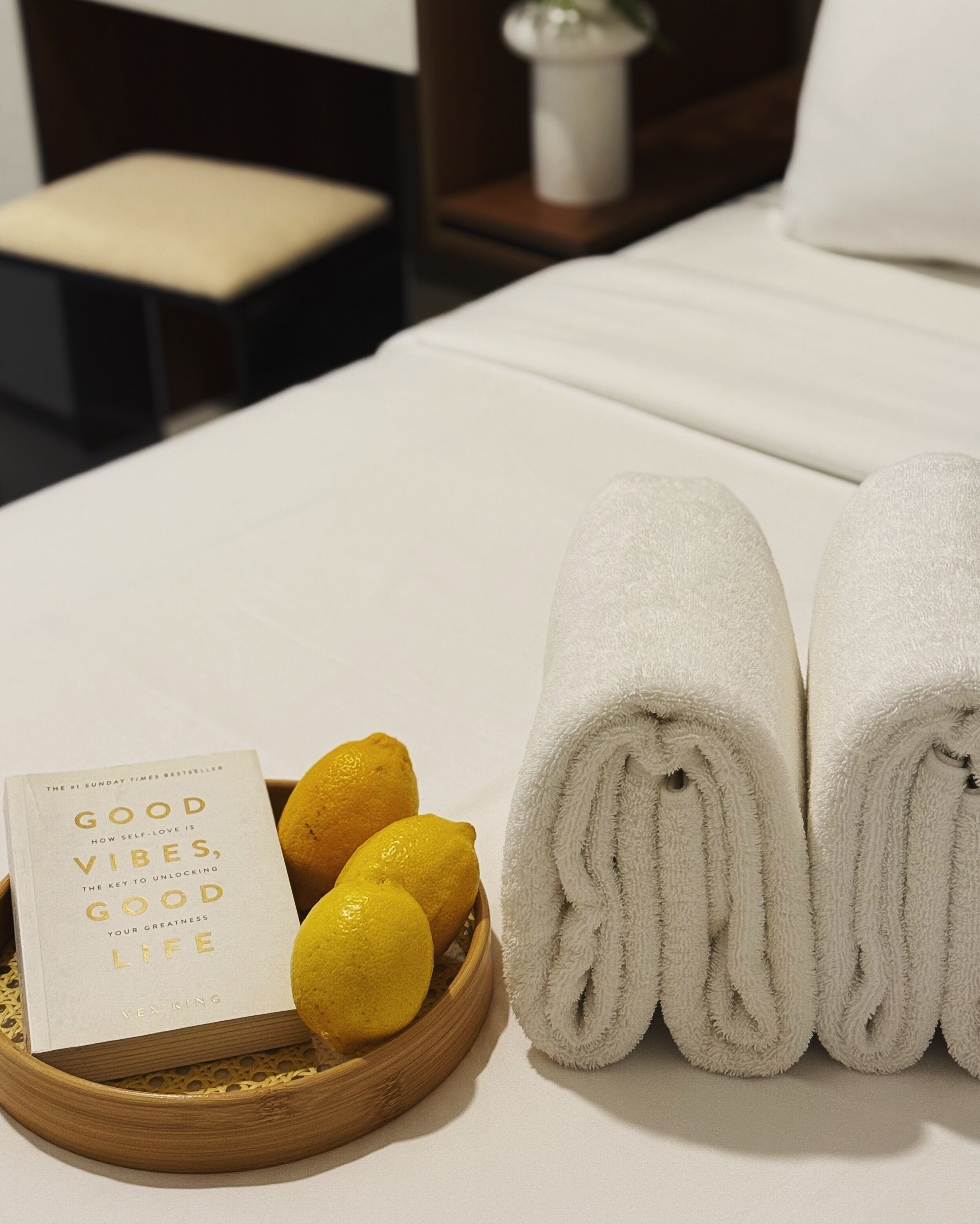 A neatly made bed with rolled white towels, a basket of lemons, and a book titled "Good Vibes, Good Life" by Vex Ring.