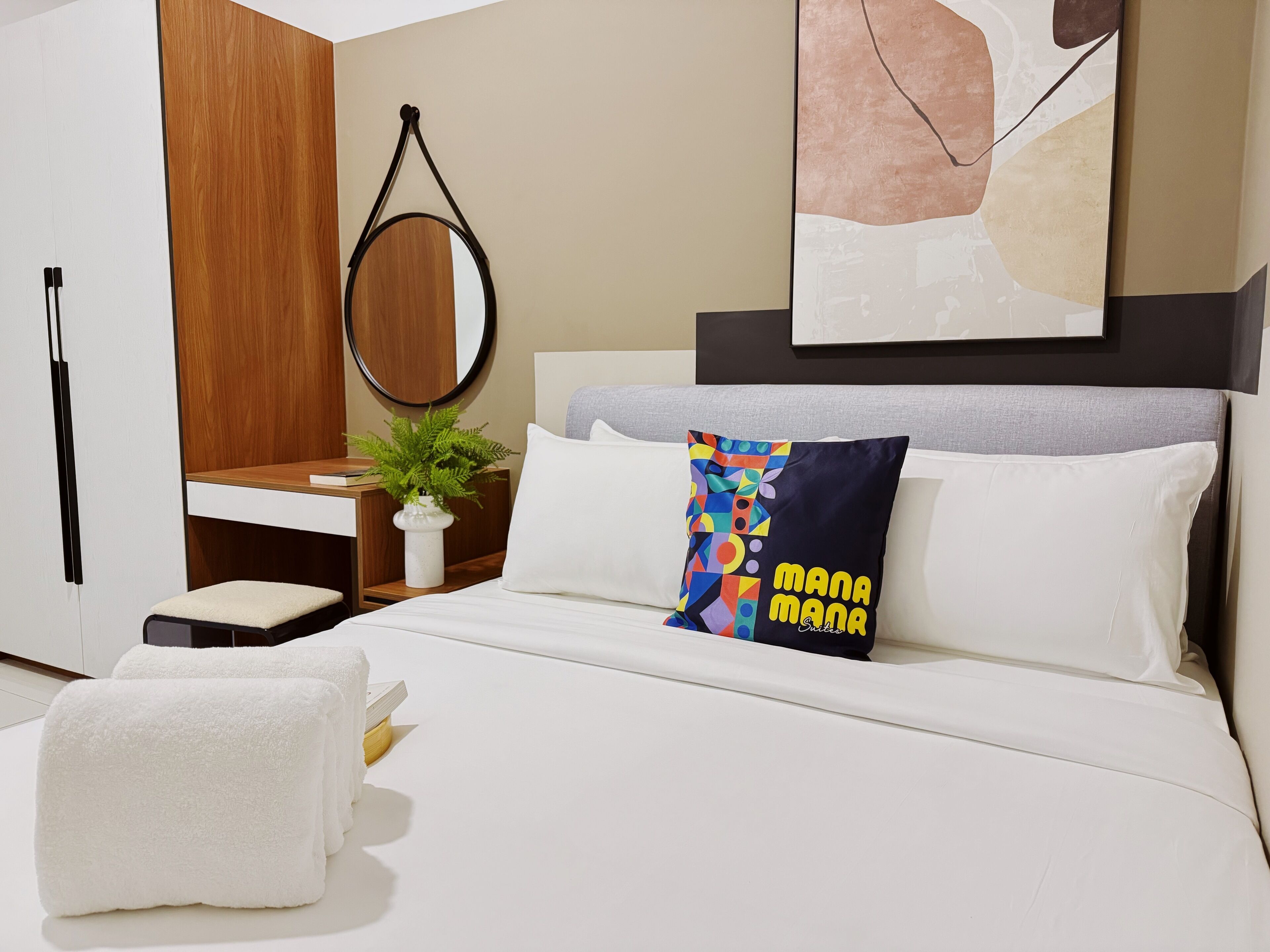 Une chambre d’hôtel moderne avec un lit, une table de chevet agrémentée d’une plante, un miroir et un coussin aux couleurs vives.