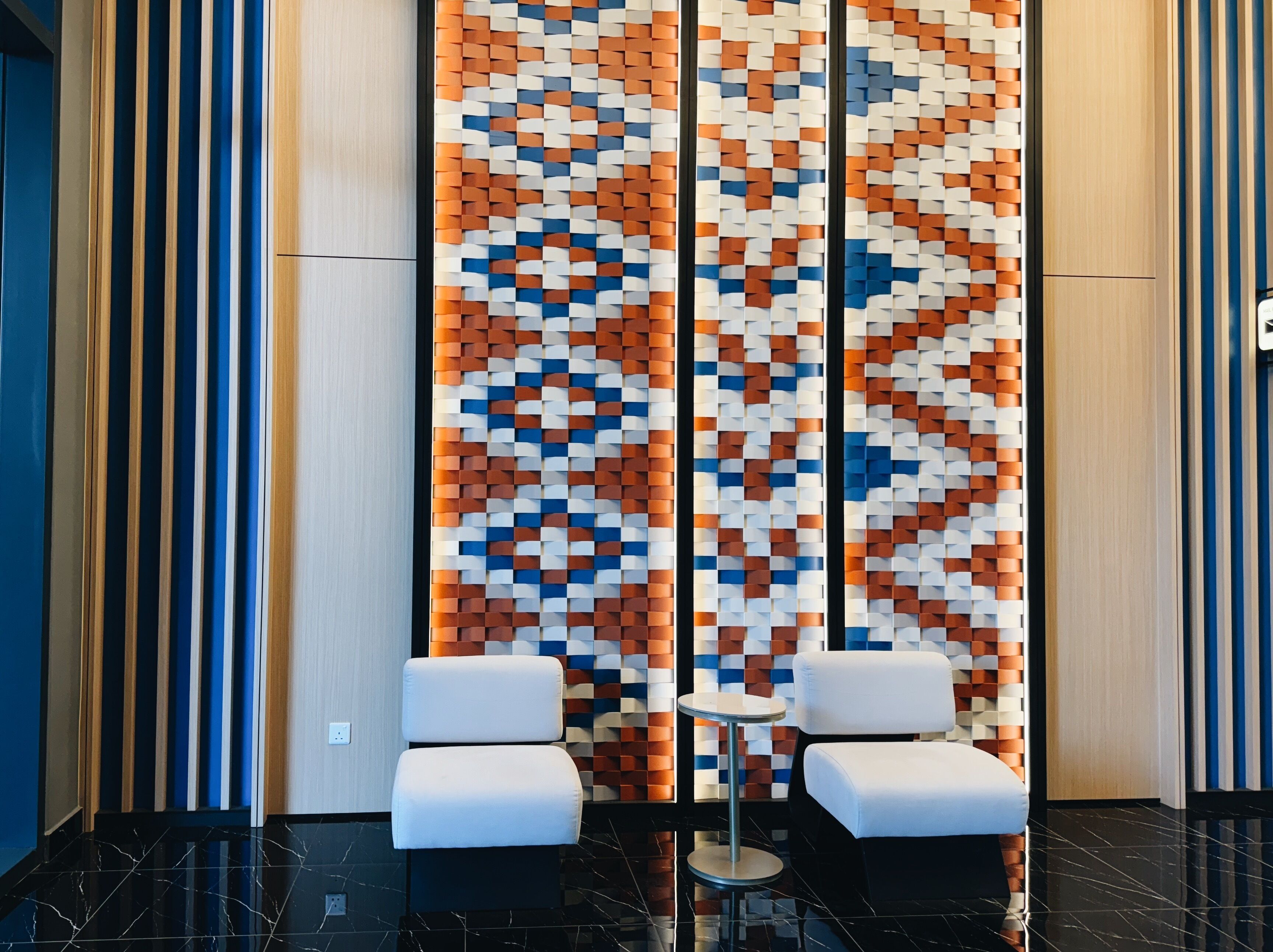 A modern lobby with a geometric patterned wall, two white cushioned seating areas, and a small round table.
