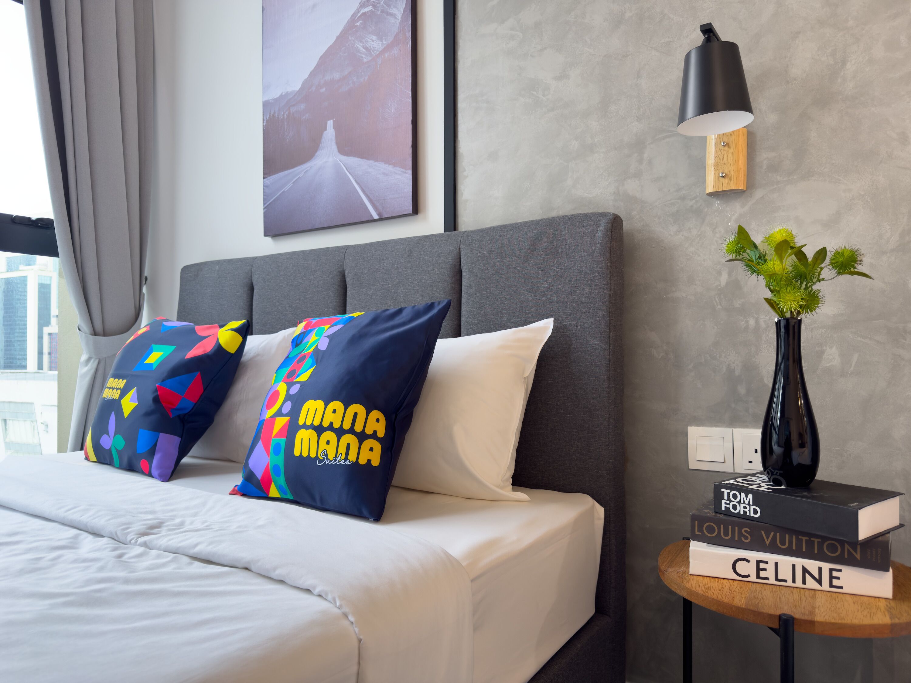 A hotel room with a bed, pillows, a nightstand with books, a vase with plants, and a wall-mounted lamp.