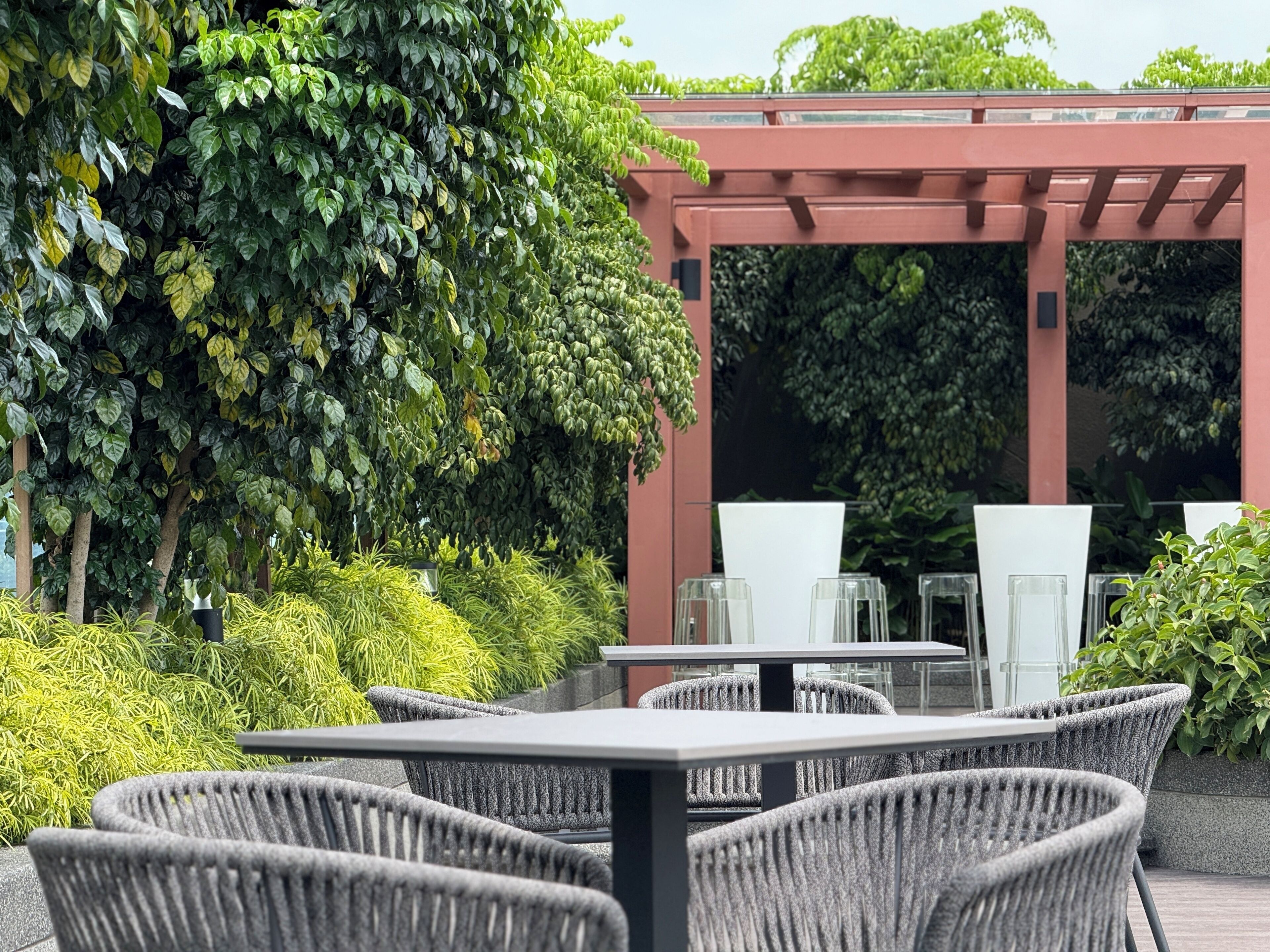 An outdoor seating area with wicker chairs and tables, surrounded by lush greenery and a red pergola structure.