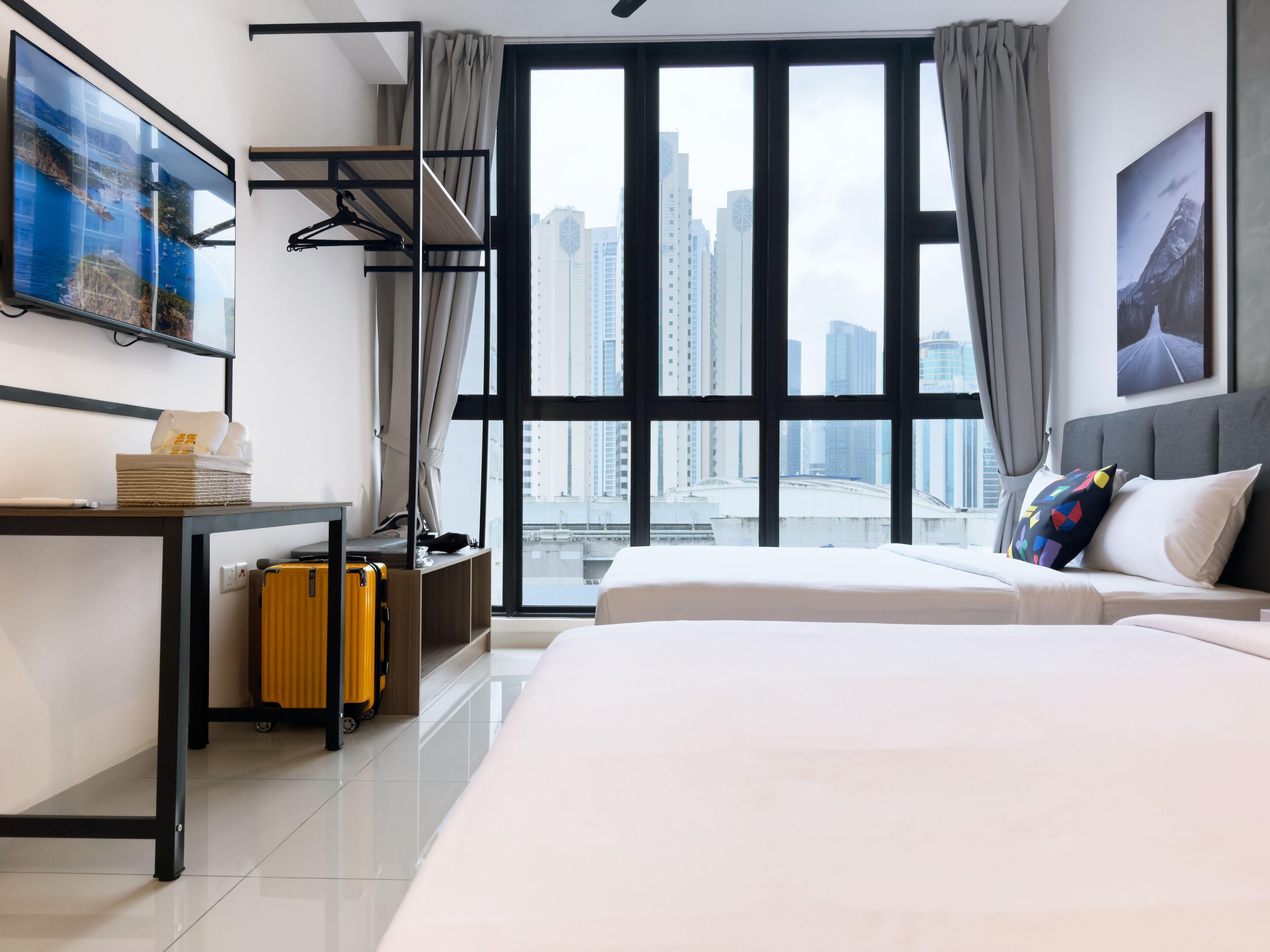 A modern hotel room with a large window, a bed, a desk with a suitcase, and a coat rack.