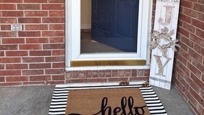 Ein Eingangsbereich eines Ziegelhauses mit einer weißen Tür, einer Fußmatte mit der Aufschrift "hello" und einem dekorativen Schild mit dem Buchstaben 'J' und einem Kranz.