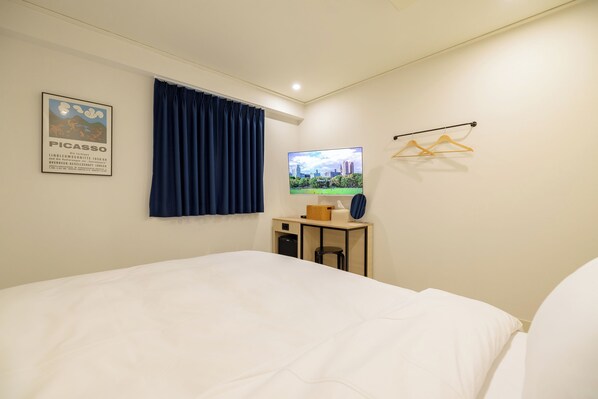 A hotel room with a bed, a desk with a computer monitor, a small table with a lamp, a window with curtains, and a poster on the wall.