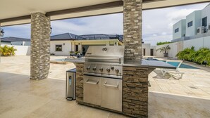 An outdoor kitchen with a built-in grill, stone pillars, and a pool area. - Stunning Luxury Pool OasisRooftop W/ Ocean Views (Noord)