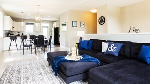 A modern living room with a dark sofa, a coffee table with a bottle and glasses, and a dining area with chairs and a table. - Bright 6 BD Executive Home - 2 Kitchens - Garage - NFLX (Edmonton)