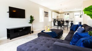 A modern living room with a large flat-screen TV, a dark grey sofa, and a dining area with black chairs. - Bright 6 BD Executive Home - 2 Kitchens - Garage - NFLX (Edmonton)