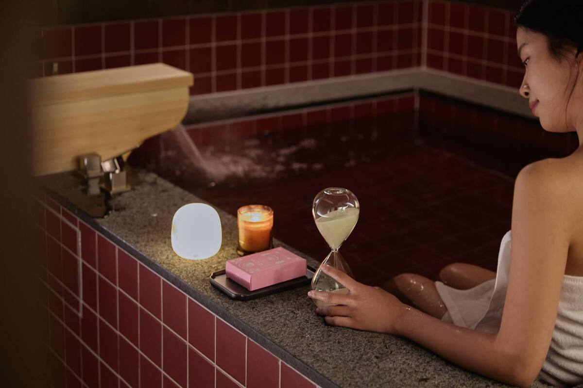 Une personne est assise près du comptoir d’une salle de bain, avec une bouteille de lotion, une bougie et un verre d’eau.