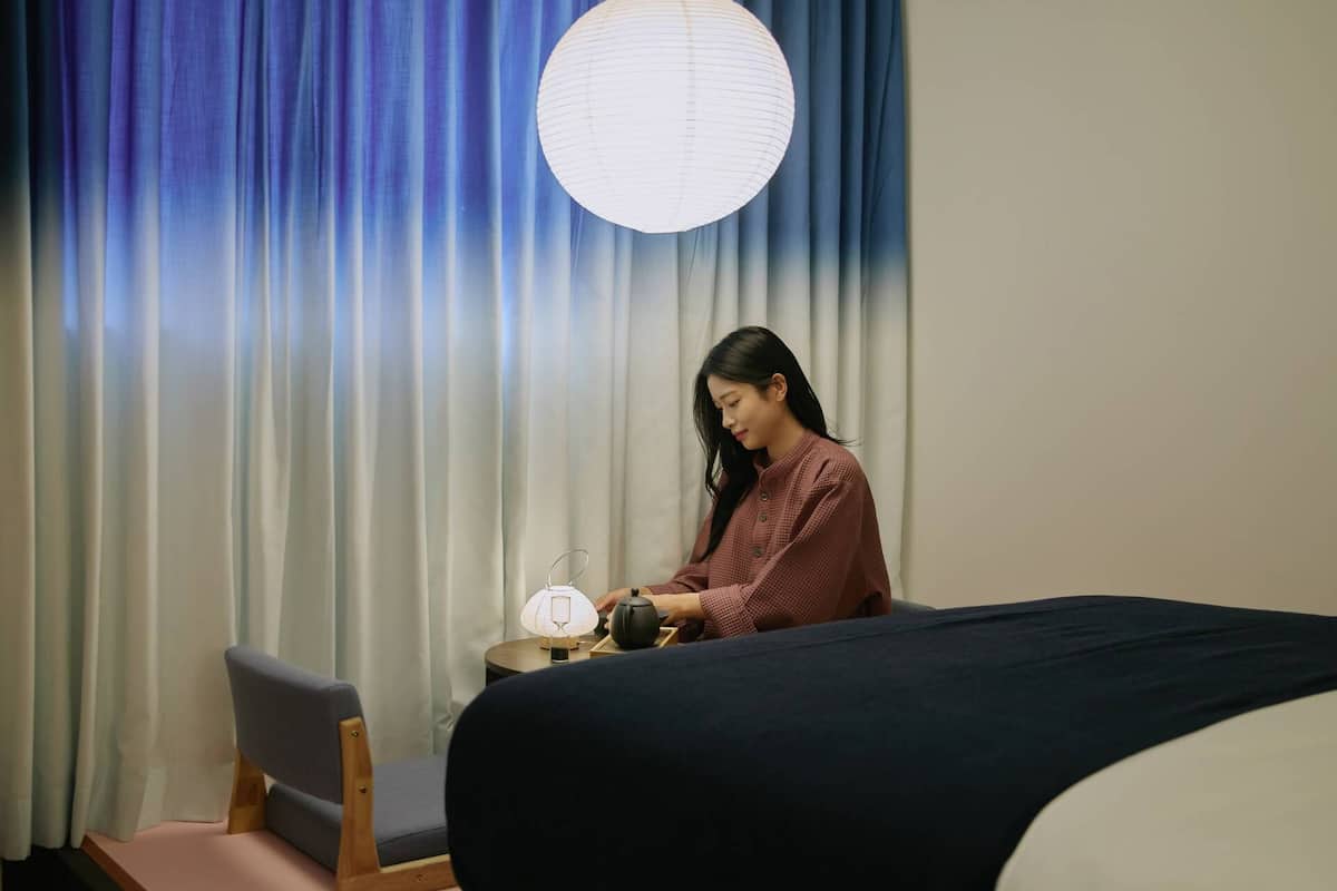 Une pièce comprenant un lit, une chaise, une table avec une lampe et une théière, ainsi qu’une lanterne en papier suspendue.