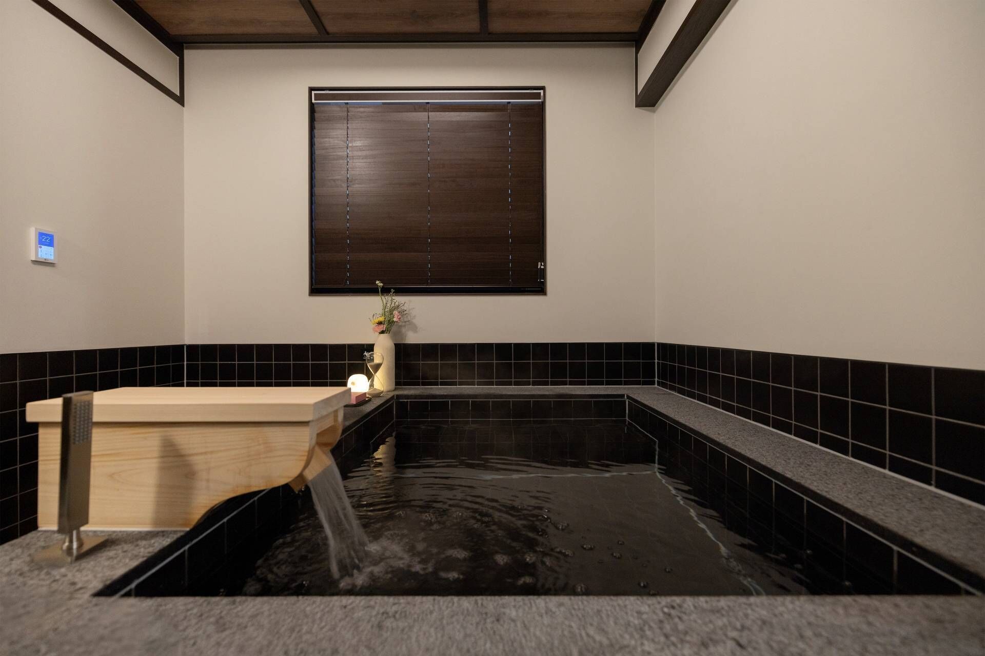 A modern bathroom with a wooden bathtub, black tiled walls, and a window with blinds.