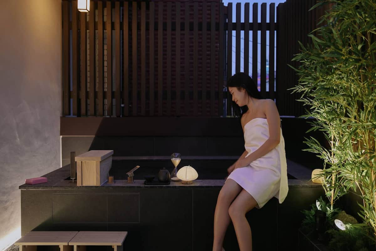 Une personne est assise sur un banc en bois, dans un espace extérieur peu éclairé, avec une petite table et une lampe.