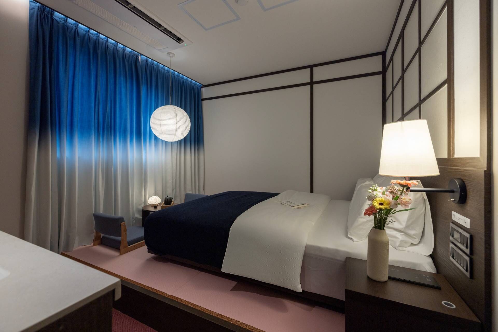 A hotel room with a bed, bedside table with a lamp and flowers, a desk, and a chair.