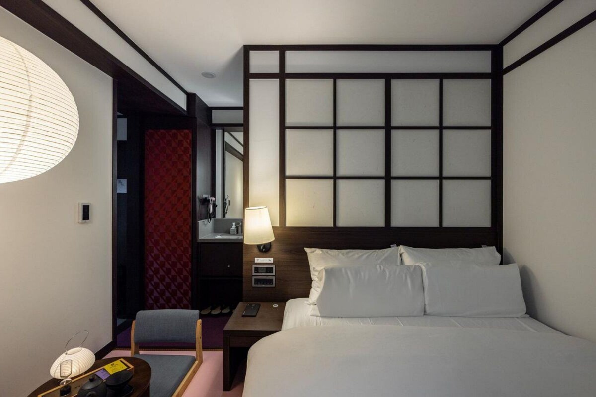 Ett hotellrum med en säng, ett sängbord, en lampa, en stol och en vägg med ett geometriskt mönster.