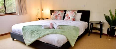 A neatly made bed with a green blanket, white pillows, and a white duvet. There is a small bedside table with a lamp, a potted plant, and a wall-mounted light fixture.