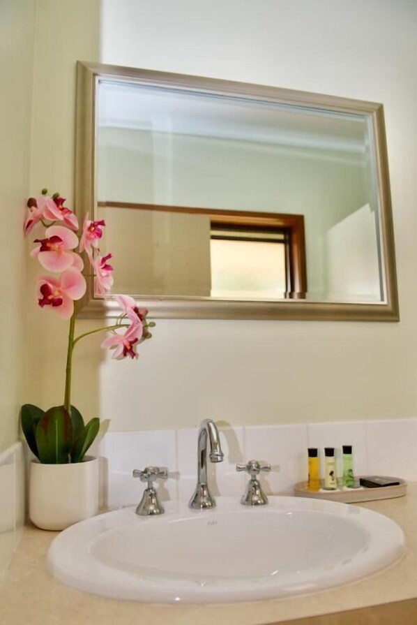 A bathroom with a white sink, two faucets, a mirror, and a potted orchid. - Green Gables Motel (Warburton)