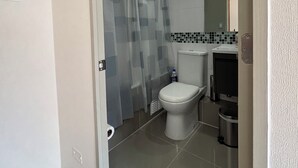 Kylpyhuone, jossa on wc, suihkuverho, laatoitettu lattia ja roskakori.