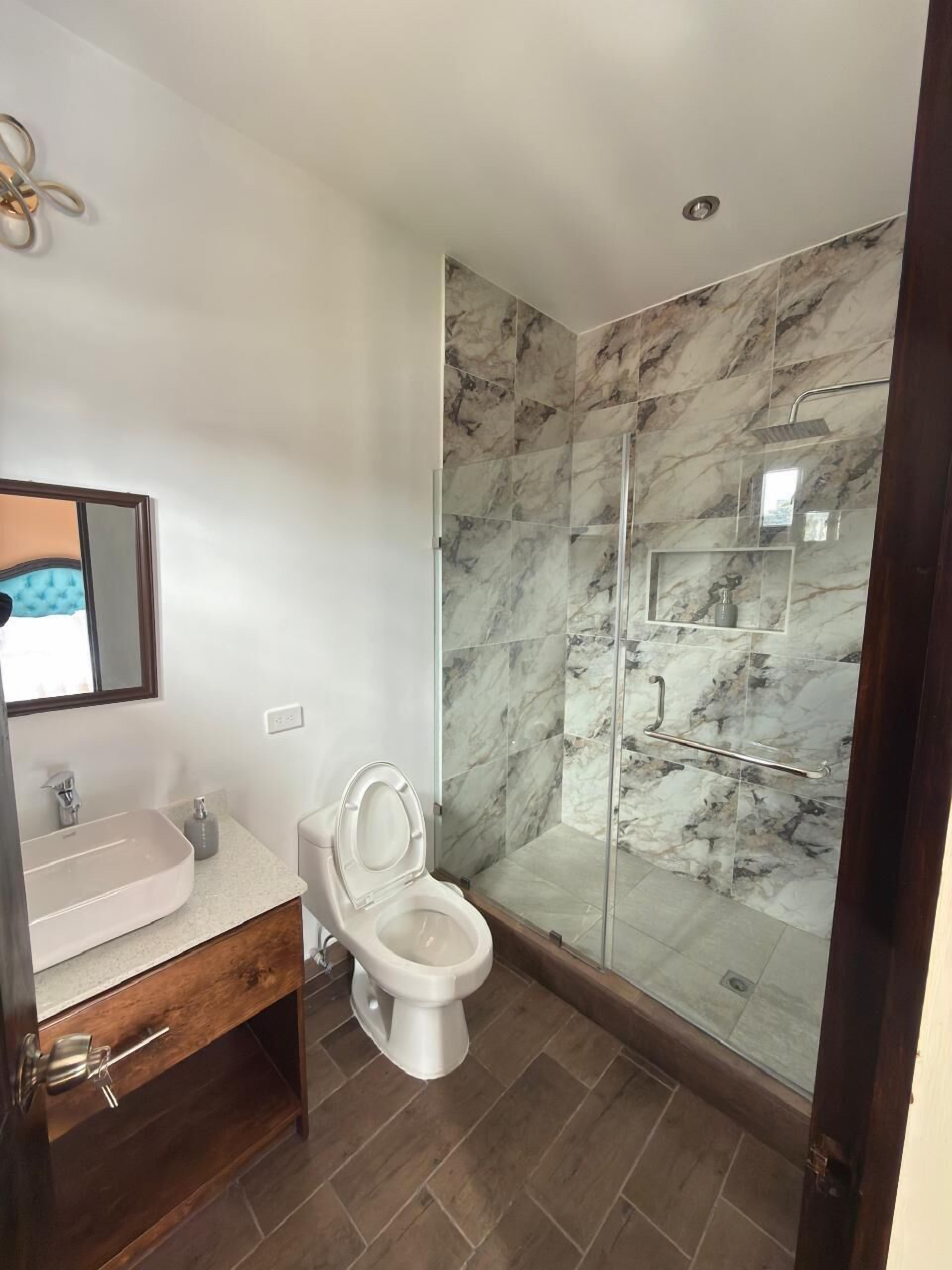 Un baño con ducha de mármol, inodoro blanco, tocador con espejo y mueble de madera.
