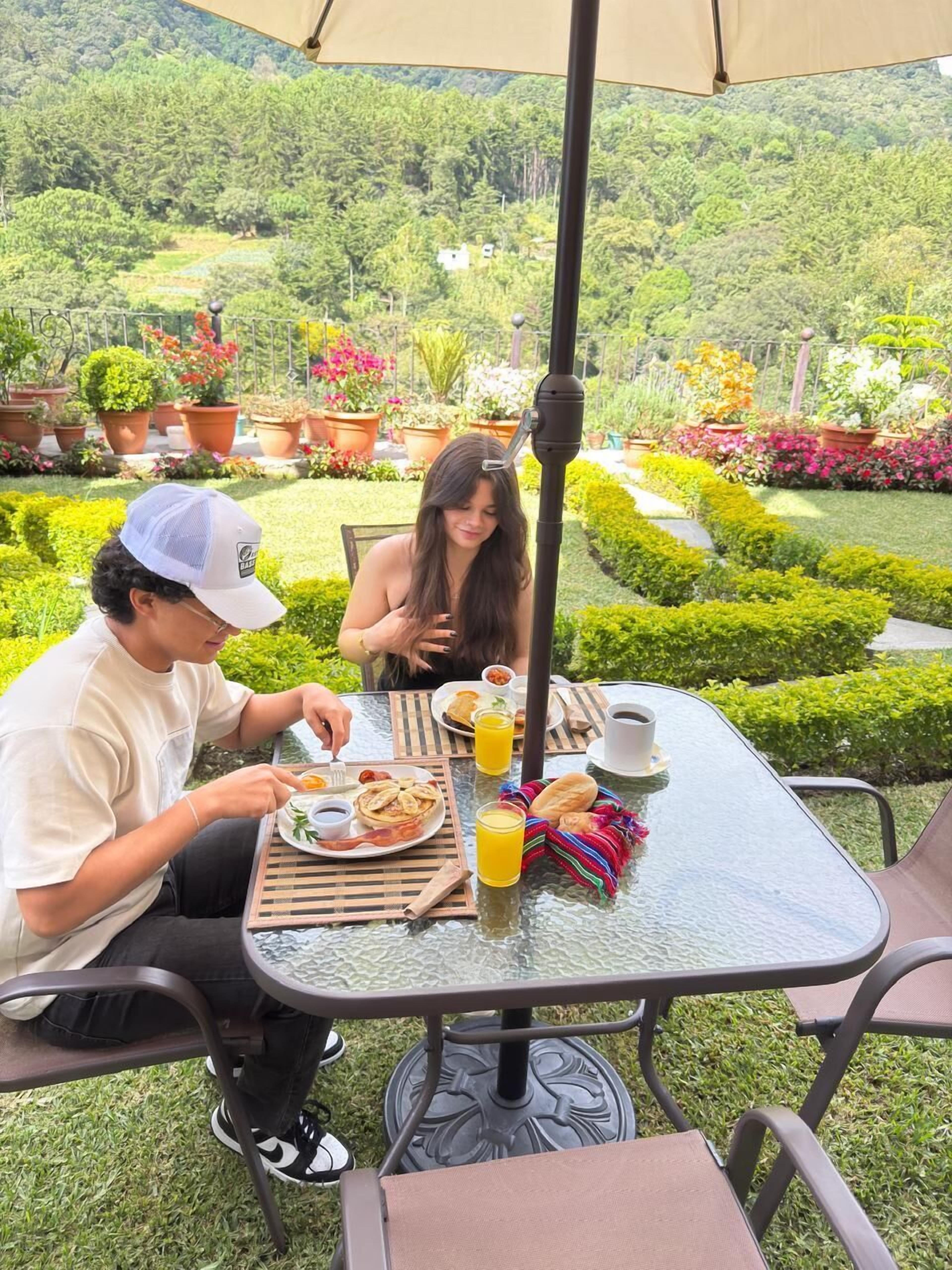 Deux personnes dînent à une table d’extérieur dont le plateau est en verre, entourées d’une végétation luxuriante et de plantes en pot.