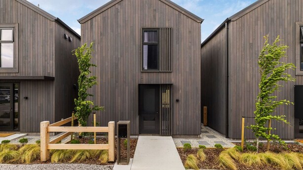 A modern building with a symmetrical facade, a central entrance, and a small garden area with a wooden fence and two young trees.