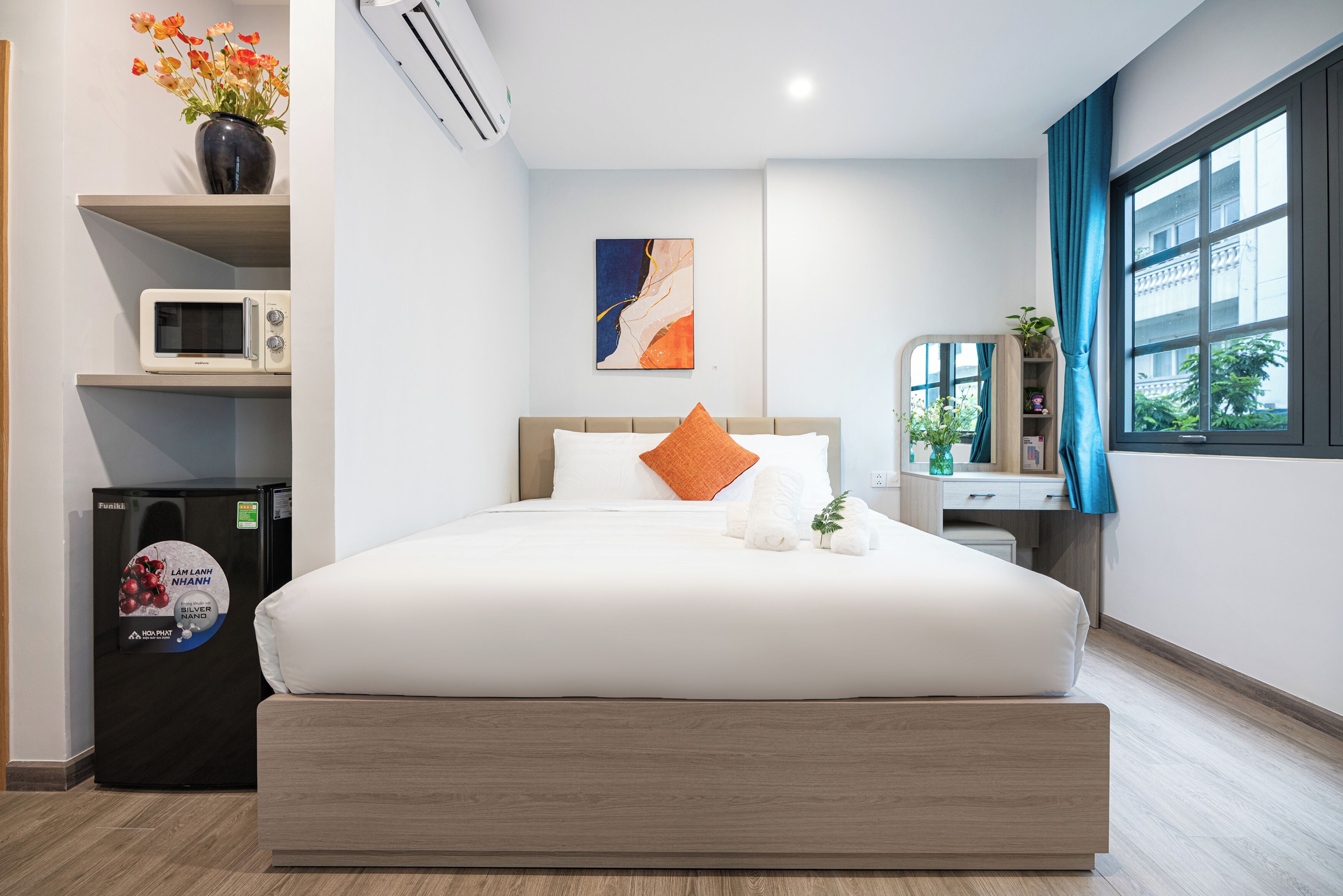 A modern hotel room with a large bed, a microwave, a refrigerator, a small plant, and a window with curtains.
