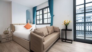 A modern hotel room with a bed, a sofa, a nightstand, a small table with a vase of flowers, and a large window with a view of buildings.
