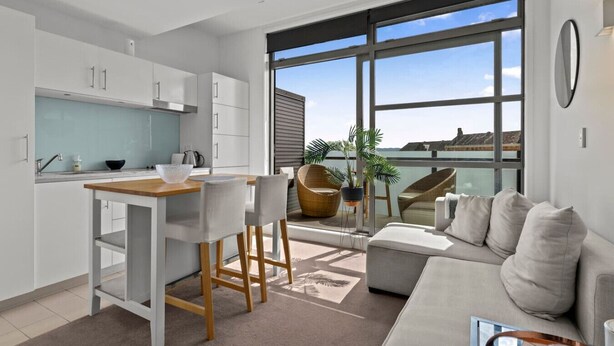 A modern kitchen with white cabinets, a wooden countertop, and a dining area with a white table and chairs. There is a large window with a view of the sea and a balcony with a sofa and chairs. - Modern Abode | Parking & Views near Ponsonby Rd (Auckland Central Suburbs)