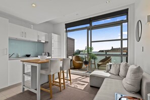 Eine moderne Küche mit weißen Schränken, einer Holzarbeitsplatte und einem Essbereich mit weißem Tisch und Stühlen. Ein großes Fenster bietet Blick auf das Meer, und ein Balkon lädt mit einer Couch und Sesseln zum Verweilen ein.