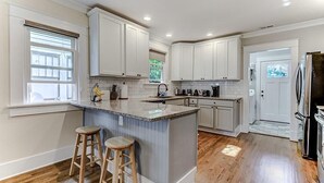 Una cocina con gabinetes blancos, una isla de granito, taburetes de madera y un refrigerador.