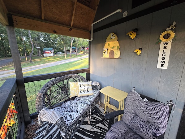 Een gezellig terras met een rieten stoel, een klein tafeltje en een bank met gestreepte kussens. Een 'Bee KIND' kussen ligt op de stoel, en een 'WELCOME' bordje hangt aan de muur.