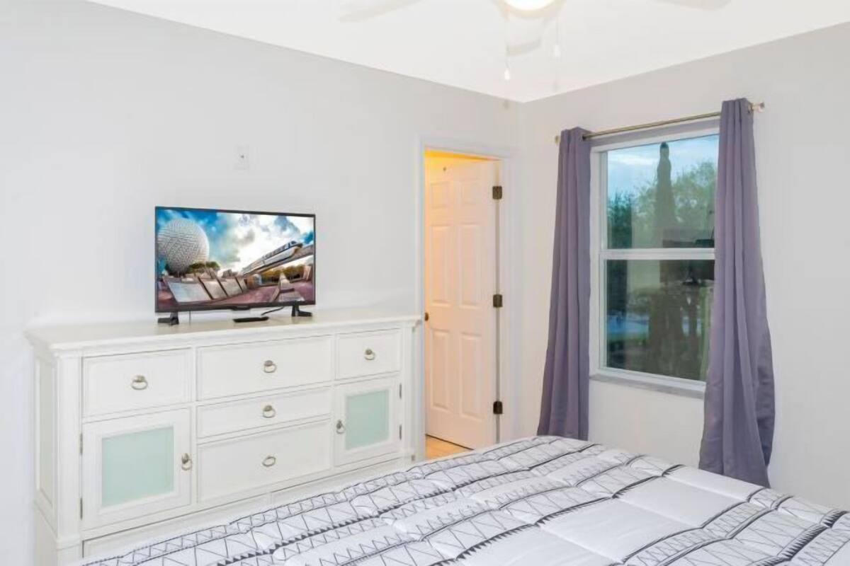 A bedroom with a bed, a dresser, a television on the dresser, a window with curtains, and a door.