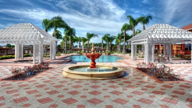 Zona de piscina con una fuente central, gazebos blancos y palmeras.