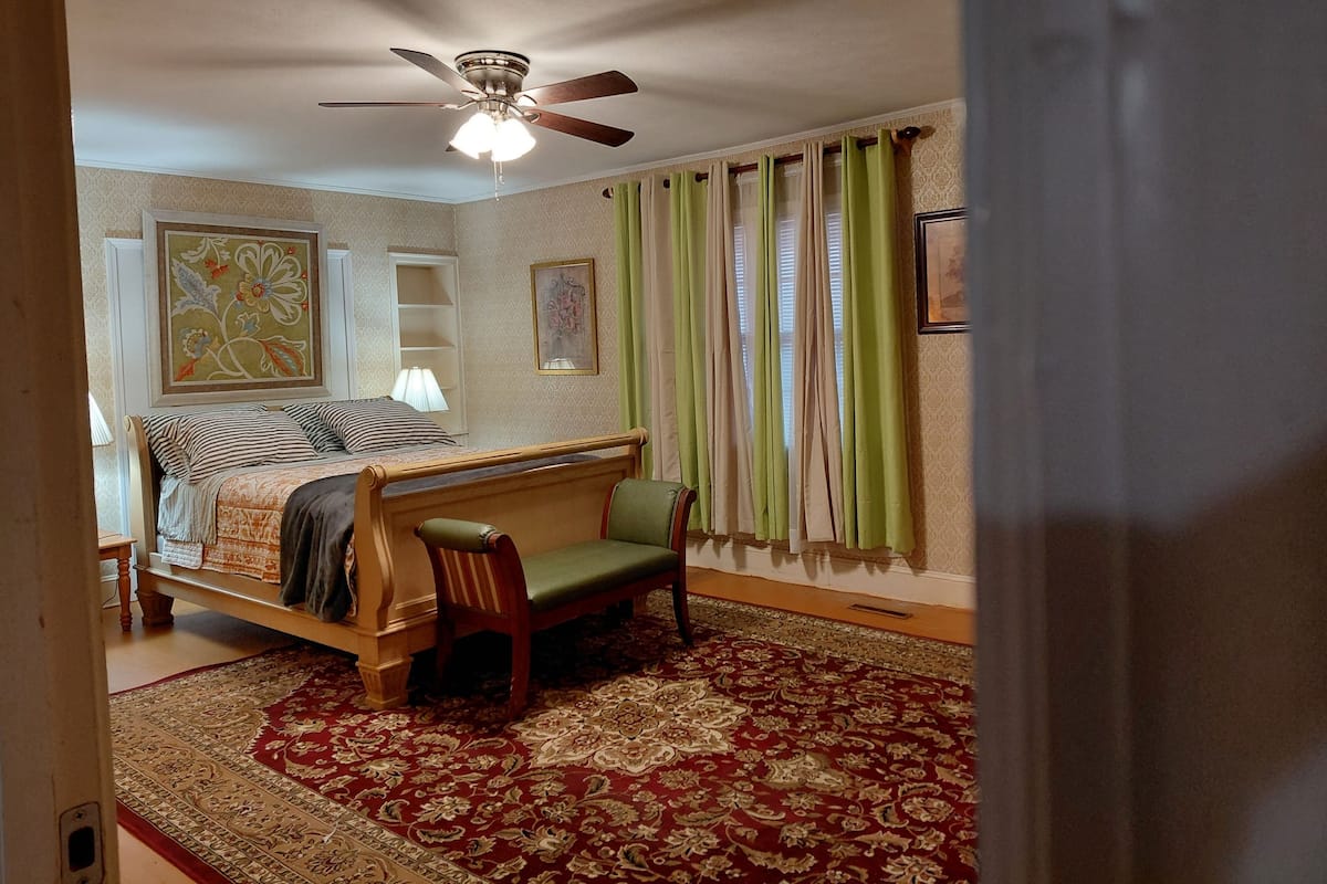 A bedroom with a bed, a chair, a ceiling fan, a window with curtains, and a patterned rug.
