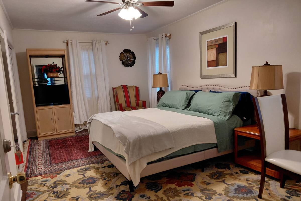 A bedroom with a bed, a chair, a desk, a ceiling fan, a window with curtains, a wall clock, and a framed picture.