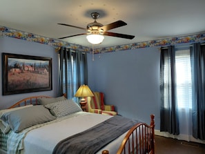 A bedroom with a bed, a ceiling fan, a lamp, a chair, a painting, and curtains.