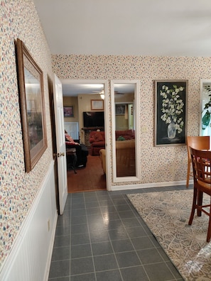A hallway with patterned wallpaper, a mirror, a dining area with a table and chairs, and a television in the background.