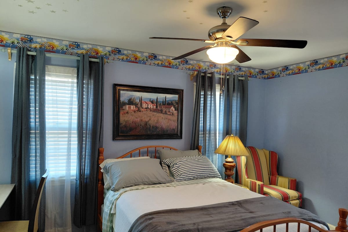 A bedroom with a bed, a ceiling fan, a painting, and curtains.
