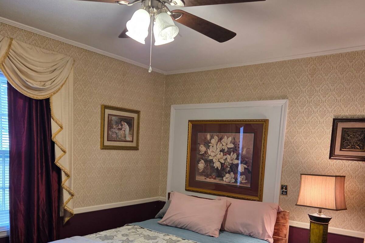 A bedroom with a bed, a ceiling fan, a nightstand with a lamp, and two framed pictures on the wall.