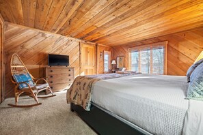 A cozy bedroom with a wooden ceiling, a bed with a quilt, a rocking chair, a dresser, and a television. - Bear Necessities | Cozy Waterfront Cabin | Lake Views, Pet Friendly, Kayak (Rangeley)