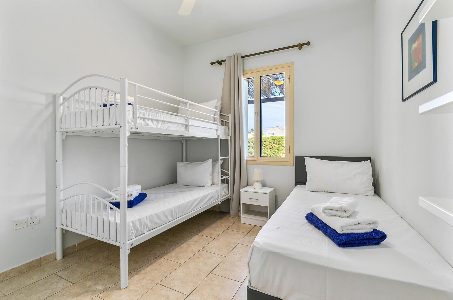 A room with a bunk bed, a single bed, a nightstand, a window with curtains, and a framed picture on the wall.
