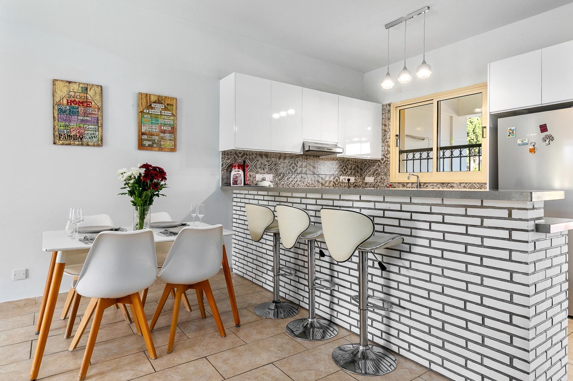 A modern kitchen with white cabinets, a brick backsplash, and a bar area with stools. There are two decorative signs on the wall and a window with a view.