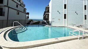 En swimmingpool med klart, blåt vand, omgivet af en hvid bygning med balkoner, og havudsigt i baggrunden.