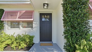 Une maison avec une porte d’entrée sombre, un petit porche avec un paillasson et une fenêtre avec des volets.