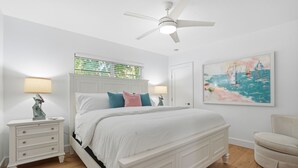 Une chambre avec un lit blanc, deux tables de chevet avec des lampes, un ventilateur de plafond, une porte et un tableau au mur.
